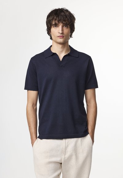 Junger Mann mit dunklem, welligem Haar, der ein marineblaues Poloshirt mit kurzen Ärmeln und hellbeige Hose trägt und mit den Händen in den Taschen steht.