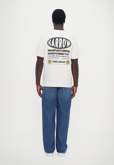 T-shirt in cotone crema con testo grafico nero sulla schiena, abbinata a jeans blu a vestibilità rilassata e sneakers bianche.