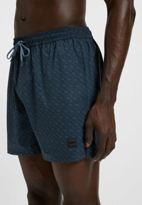 Dunkelhäutiger Mann in navy-blauen gemusterten Badeshorts mit Kordelzug, steht vor einem schlichten hellgrauen Hintergrund, die rechte Hand entspannt an seiner Seite.