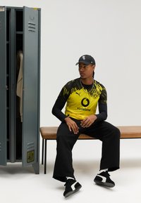 Geltonas ir juodas „Puma“ futbolo marškinėlių komplektas su „Vodafone“ logotipu, po juo juoda ilgomis rankovėmis marškinėliai, juodos kelnės ir juodi sportbačiai. Fone matoma pilka spintelė.