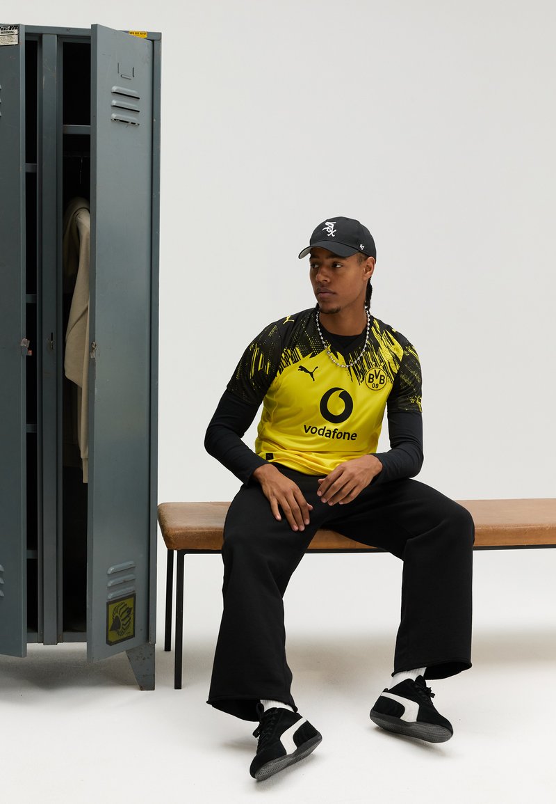 Žlutý a černý fotbalový dres Puma s logem Vodafone, pod ním černé tričko s dlouhým rukávem, černé kalhoty a černé tenisky. V pozadí šedá skříňka.