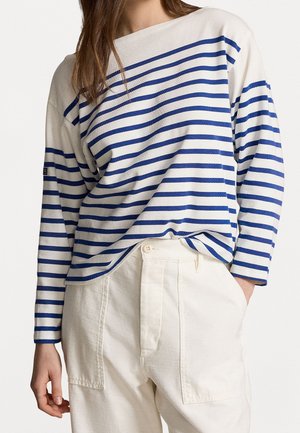 Personne portant un haut blanc à manches longues avec des rayures horizontales bleues et un pantalon blanc taille haute avec des poches plaquées à l'avant.