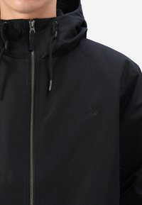 Juoda zip-up striukė su kapišonu, pasižyminti glotnia tekstūra, matomu užtrauktuku ir subtiliu logotipo akcentu ant krūtinės.