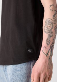 Tricou negru din bumbac cu un design ajustat, având un mic etichet cu logo-ul Calvin Klein la tiv și un tatuaj detaliat cu un șarpe pe braț.