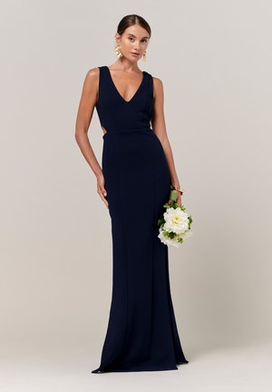 Frau in einem ärmellosen marineblauen Kleid, die weiße Blumen hält, goldene Ohrringe und ein Armband trägt und vor einem einfarbigen Hintergrund steht.