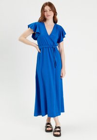 Robe enveloppante bleue avec des manches flottantes, fabriquée à partir d'un tissu texturé. La robe présente une taille cintrée et tombe jusqu'aux chevilles, accompagnée de sandales noires.