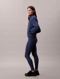 Sinine tõmblukuga jakk kõrge kraega, koos siniste legging'itega, millel on diagonaalne õmblus. Mustad spordijalatsid täiendavad komplekti.