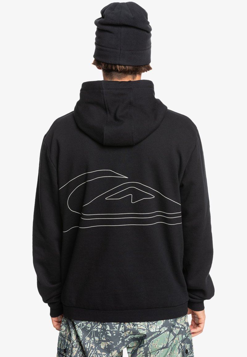 Crna dukserica s velikim bijelim obrubom grafike na leđima. Ima kapuljaču i rebraste manžetne. Model nosi crnu beanie i šarene hlače.