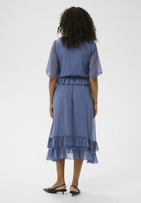 Vestido azul claro, semitransparente, con bajo con volantes y mangas cortas, presenta una cintura ajustada y una silueta fluida. Zapatos de tacón negro con tira en el talón.