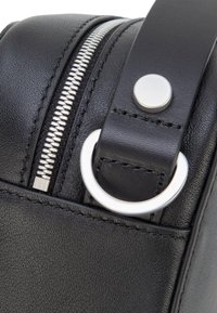 Detailní záběr černé kožené tašky, zobrazující stříbrný zip, prošitý popruh s kulatým kovovým knoflíkem a stříbrný D-kroužek.