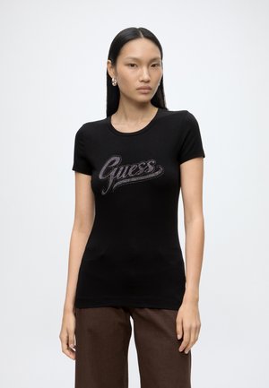Femme aux longs cheveux noirs porte un t-shirt noir Guess avec un logo en strass et un pantalon marron, debout devant un fond blanc.