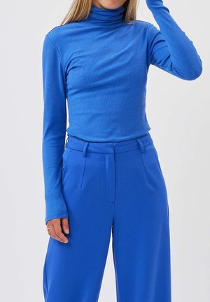 Osoba nosící jasně modrý žebrovaný rolák a modré kalhoty s vysokým pasem a širokými nohavicemi, stojící na jednoduchém bílém pozadí.