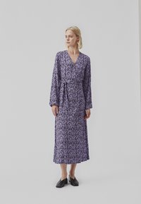 Lila blommig omlottkjol med långa ärmar, med en v-ringning och knytbart midjeband. Slätt tyg med små vita blommönster.