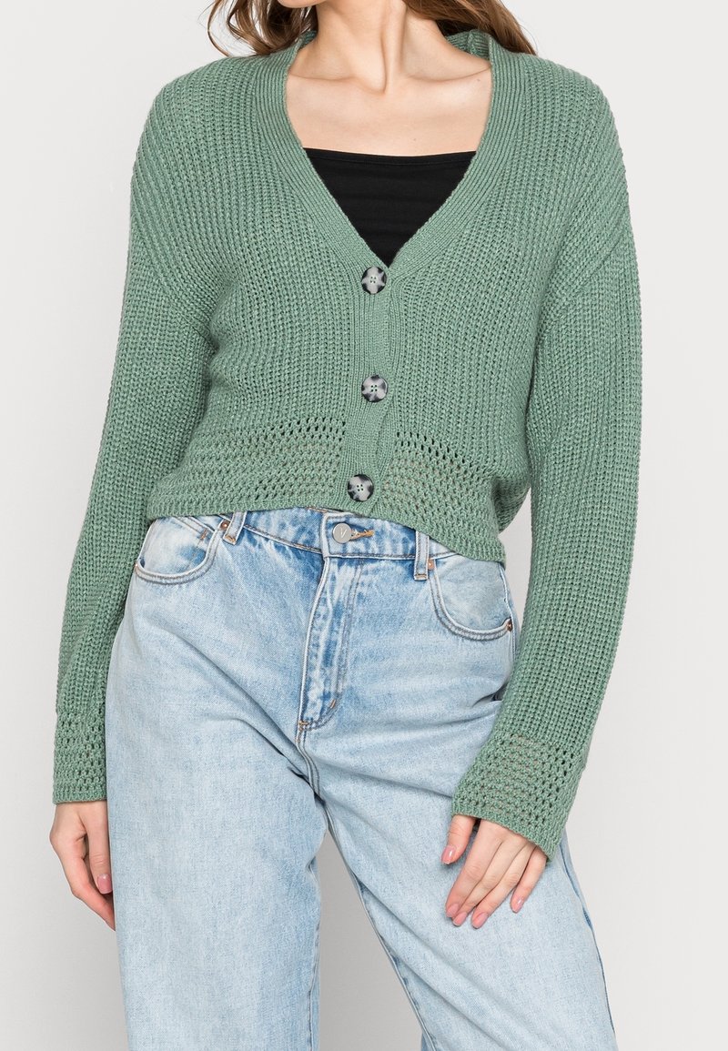 Cardigan en tricot vert avec un design court, présentant un décolleté en V profond, six boutons noirs et des poignets à côtes. Porté sur un haut noir et un jean clair.