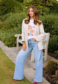 Beige Blazer über einem weißen Grafik-T-Shirt mit einem Getränk und buntem Design, kombiniert mit hellblauen weiten Jeans und roten Schuhen.