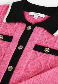 Cardigan tricoté rose avec col et bordures noires, boutons dorés texturés et une étiquette blanche portant l'inscription "NEXT" à l'intérieur du col.