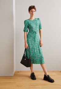 Zielona kwiatowa sukienka midi z krótkimi bufkami, taliowana paskiem i o zwiewnym kroju. W połączeniu z czarnymi mokasynami na obcasie i czarną torebką.