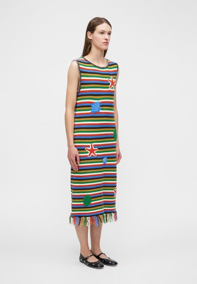 Frau steht in einem bunten, gestreiften, ärmellosen Strickkleid mit Seestern- und Muschelmustern und Fransen am Saum, trägt schwarze Ballerinas.