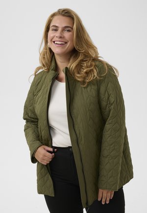 Femme souriante portant une veste matelassée vert olive par-dessus un haut blanc et un pantalon noir sur un fond clair uni.
