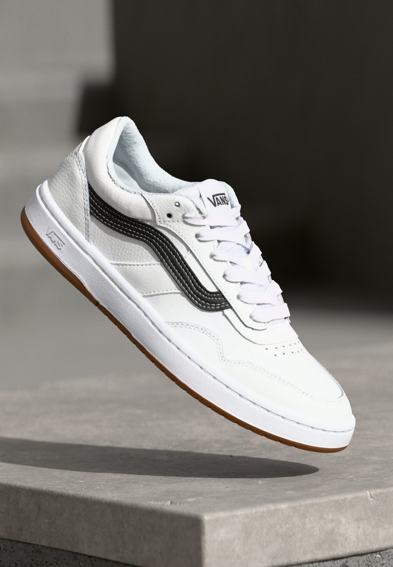 Baskets blanches Vans avec bande noire et semelle en gomme au-dessus d’un rebord en béton sous un éclairage doux.