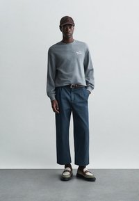 Sivá dlhá mikina s bielym logom, tmavomodré široké nohavice a hnedé a biele topánky. Neformálny dizajn s voľným strihom.