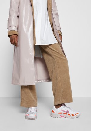Personne portant un pantalon beige en velours côtelé, une chemise blanche, un trench-coat beige brillant et des baskets blanches avec des touches orange et rose, debout devant un fond uni.