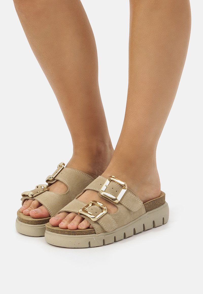 Le ciabatte in suede beige presentano doppie fibbie regolabili, un fondo in sughero e una suola in gomma spessa e testurizzata con scanalature orizzontali.