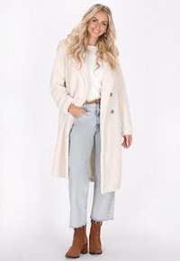 Krémový kabát z umělé kožešiny s oversize střihem, dvěma předními knoflíky a bočními kapsami, v kombinaci se světle modrými džínami s vysokým pasem a hnědými kotníkovými botami.