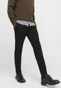 Czarne, dopasowane spodnie wykonane z gładkiego materiału z bocznymi kieszeniami, noszone z brązowym swetrem i pod spodem pasiastą koszulą.