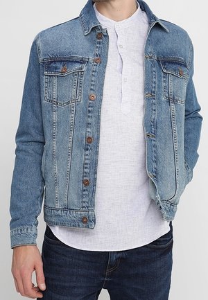 Mann trägt eine hellblaue Jeansjacke über einem weißen Henley-Hemd mit Knöpfen und dunkelblaue Jeans, steht mit linker Hand in der Tasche.