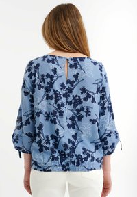 Blusa floral azul con patrones en navy, mangas tres cuartos, espalda con abertura en forma de llave y cierre de botón, y dobladillo elástico. Textura de tela ligera.