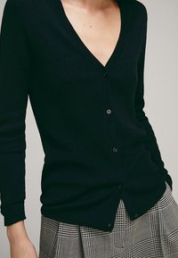 Cardigan noir côtelé avec col en V et manches longues, doté de boutons. Porté avec une jupe grise à carreaux, présentant des motifs distincts.