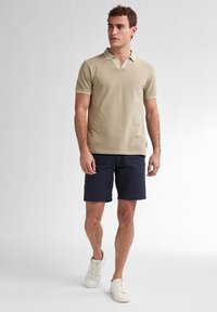 Béžové polo tričko s límečkem, spárované s tmavě modrými šortkami. Model má na sobě bílé tenisky. Hladká textura a volný střih.