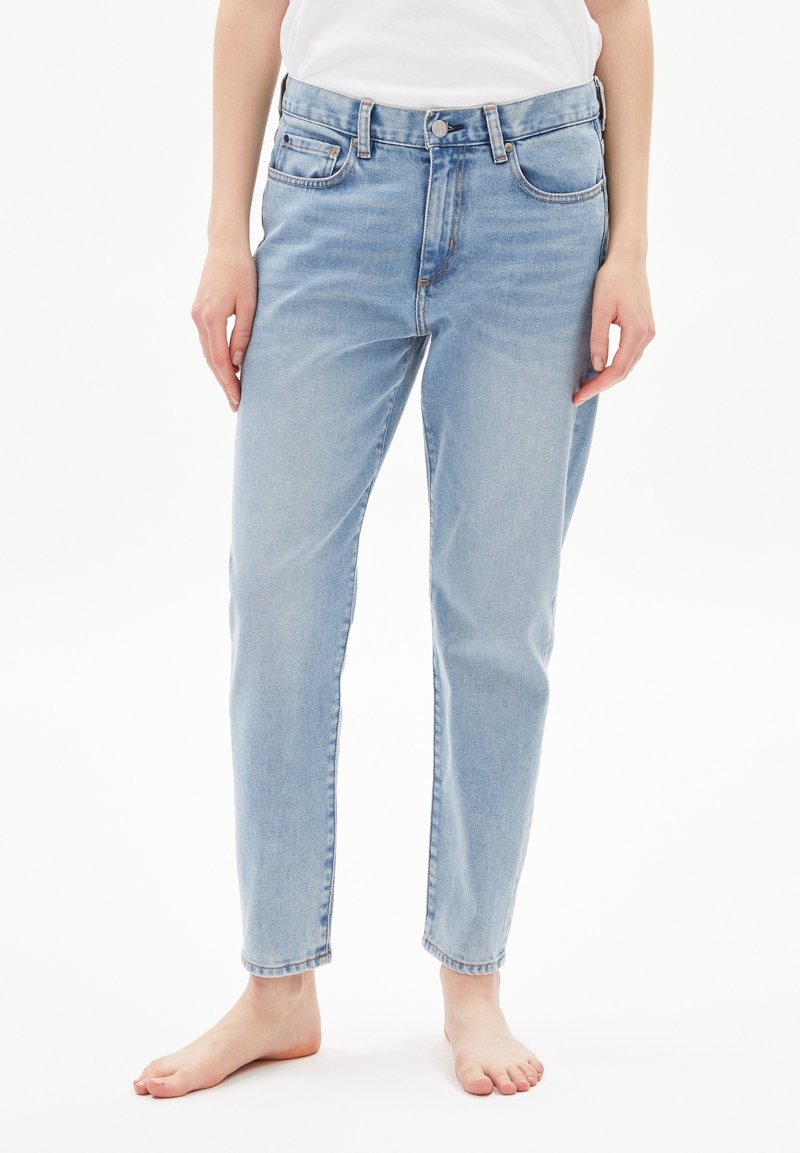 Personne portant un jean en denim bleu clair et une chemise blanche, debout pieds nus contre un fond blanc.