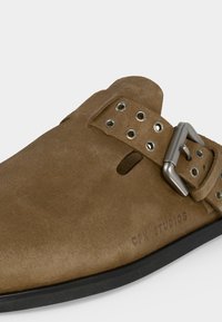 Καφέ σουέτ παπούτσι slip-on με ρυθμιζόμενο μεταλλικό κούμπωμα και πολλαπλές τρύπες στο λουρί, μαύρη σόλα και ανάγλυφο "GHI STUDIOS" στο πλάι.