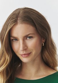Nahaufnahme einer Person mit langen, welligen braunen Haaren, die ein grünes Kleidungsstück und goldene Creolen trägt. Die Person hat einen glatten Teint und blaue Augen.