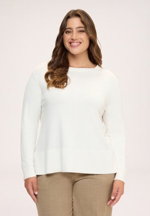 Femme souriante aux cheveux bruns ondulés portant un pull blanc à manches longues et un pantalon beige, debout devant un fond clair.