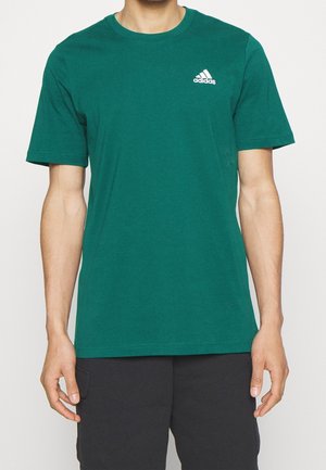 Mężczyzna w zielonym t-shircie Adidas z białym logo na piersi i czarnych szortach, stojący na jednolitym tle.
