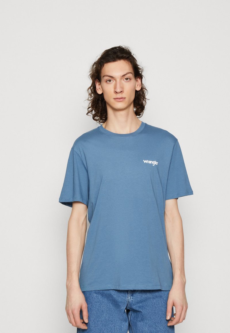 T-shirt in cotone blu con maniche corte, scollatura rotonda e logo bianco "Wrangler" sul lato sinistro del petto. Indossato con jeans in denim blu.