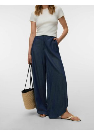 Žena v bielom tričku s krátkym rukávom, široké tmavomodré nohavice, béžové sandále, drží slamennú tašku s čiernymi rúčkami.