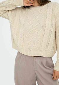 Beiger Strickpullover mit Zopfmuster, gerippten Bündchen und Saum; kombiniert mit hellbraunen, plissierten Hosen. Glatte Textur, entspannte Passform.