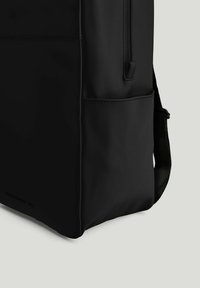 Sort rygsæk hjørne med sidelomme, lodret lynlås og polstret skulderrem mod en lys baggrund.