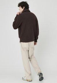 Pull marron à côtes avec un col montant, associé à un pantalon beige clair à coupe décontractée et des baskets multicolores. Design minimaliste, look décontracté.