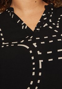 Svart plagg med V-ringning och ett mönster av vita streckade linjer och geometriska former på tyget, med en slät yta.