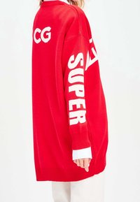 Rode oversized trui met witte "SUPER" tekst op de mouwen en "CG" op de achterkant, gemaakt van zacht gebreid materiaal met geribbelde manchetten.