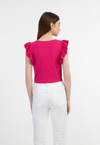 Fukszia cropped top fodros ujjakkal, texturált anyagból és kerek nyakkivágással, magas derekú fehér farmernadrággal párosítva, hátulról nézve.