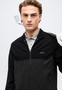 Jeune homme portant une veste noire BOSS, tenant un club de golf sur l'épaule, vêtu d'un polo gris et d'un gant de golf blanc à une main.