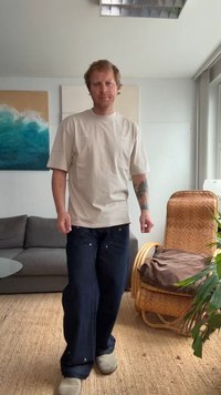 T-shirt bege de mangas curtas e jeans claros de corte largo. A modelo está em pé sobre um tapete de palha, perto de um sofá cinza e uma cadeira de vime.