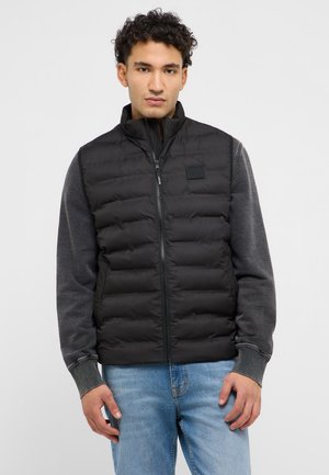 Young man wearing a black puffer vest over a dark gray sweatshirt and light blue jeans, standing against a plain light background.