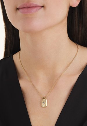 Gouden rechthoekige hangerketting met een blauw oogontwerp gedragen door een vrouw met donker haar en een zwart shirt.